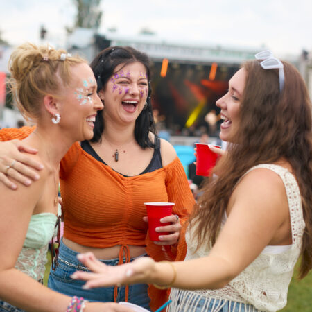 Three Female Friends Wearing Glitter Having Fun At Summer Music Festival Holding Drinks